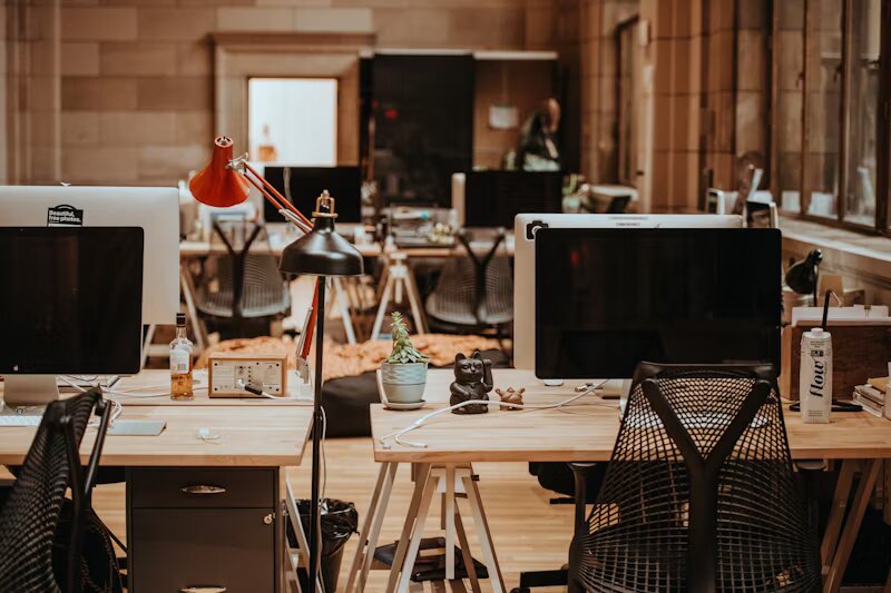 Oficina de coworking con un diseño industrial y acogedor en Spain, equipada con escritorios de madera, sillas ergonómicas, lámparas de escritorio, computadoras y decoración como plantas y figuras. El ambiente es cálido, con iluminación tenue y paredes de piedra, creando un espacio de trabajo inspirador y colaborativo.