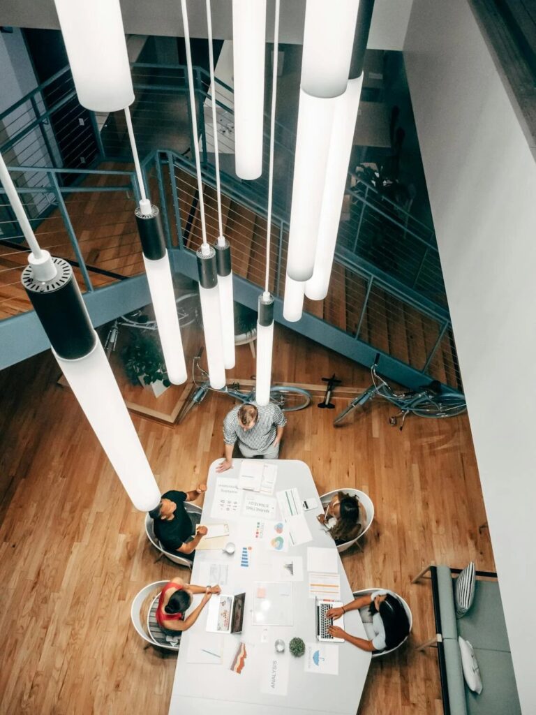 Vista aérea de compañeros de trabajo reunidos alrededor de una mesa trabajando en una sesión de estrategia bajo luces colgantes modernas.