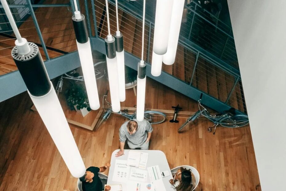 Vista aérea de compañeros de trabajo reunidos alrededor de una mesa trabajando en una sesión de estrategia bajo luces colgantes modernas.